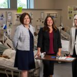 Three professionals stand in a medical training facility beside hospital beds, with one patient resting in a bed and educational posters visible on the walls.