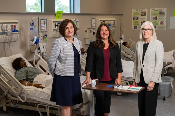Three professionals stand in a medical training facility beside hospital beds, with one patient resting in a bed and educational posters visible on the walls.