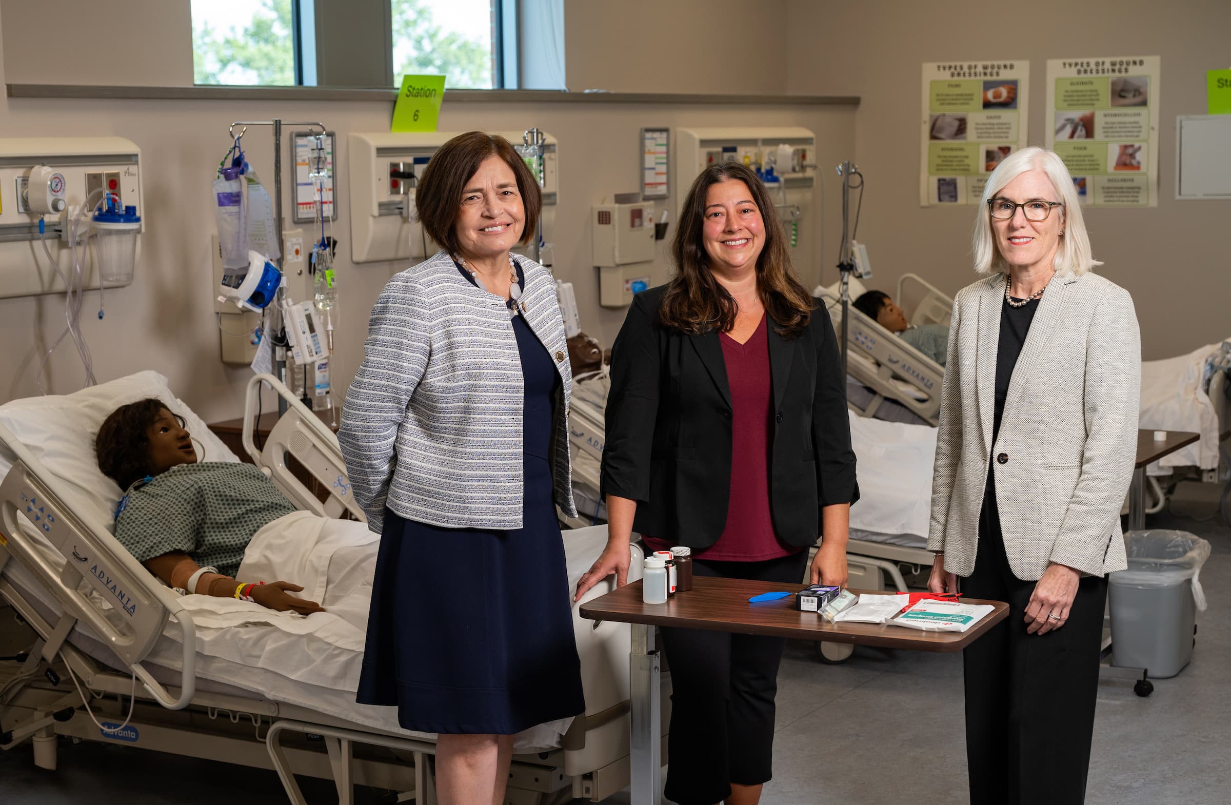 Three professionals stand in a medical training facility beside hospital beds, with one patient resting in a bed and educational posters visible on the walls.